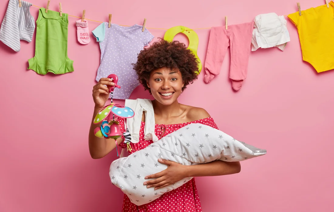 smiling-pleased-ethnic-mother-shows-mobile-toy-her-small-infant-plays-with-newborn-son-happy-become-mom-stands-indoor-against-pink-wall-baby-mums-arms-child-care-concept